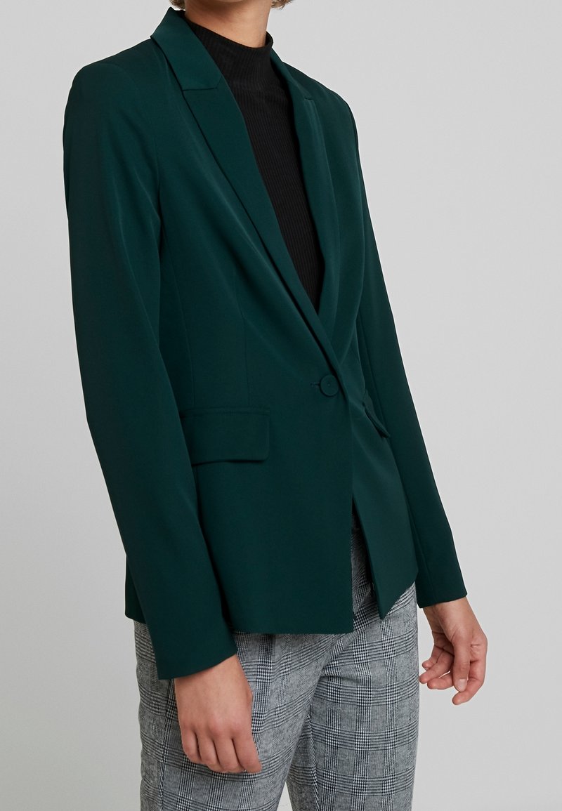 Žena v tmavě zeleném přiléhavém saku přes černý top a šedé kostkované kalhoty, stojící proti jednoduchému světlému pozadí.