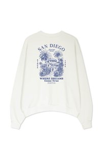 Biela mikina s modrou grafikou budovy s palmami. Text obsahuje "San Diego," "Kaotiko Resort," a "Kde sa sny stávajú skutočnosťou."