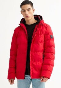 Joven con chaqueta acolchada roja con capucha, camisa negra y vaqueros azul claro, de pie frente a un fondo blanco liso.