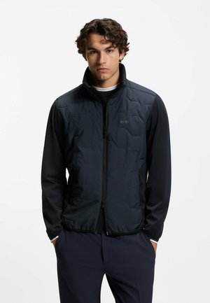 Młody mężczyzna o kręconych włosach, ubrany w czarną kurtkę na suwak z pikowanym przodem, stoi z rękami w kieszeniach na białym tle.