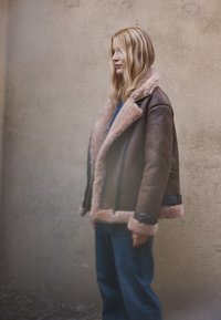 Bruine shearling jas met een tan bontkraag, zwarte accenten en brede manchetten, gecombineerd met een blauwe spijkerbroek. Tegen een textuurmuur geplaatst.