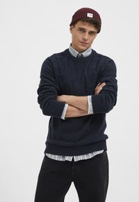Suéter de punto azul marino con cuello redondo acanalado, sobre una camisa de botones a rayas en gris claro y blanco. Gorro acanalado color burdeos en la cabeza.