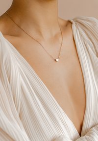Collar dorado con un pequeño colgante en forma de corazón, cadena delicada y un diseño minimalista, llevado sobre un vestido blanco texturizado.