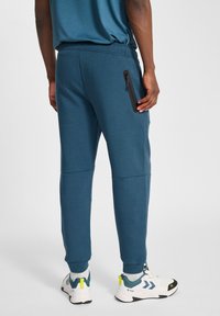 Teal joggings met een taps toelopend ontwerp, elastische tailleband en een ritssluiting zijzak. Gecombineerd met sportieve zwart-witte schoenen.