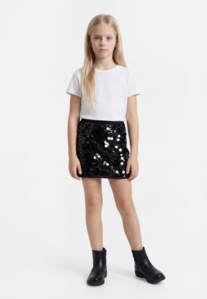 Νεαρή κοπέλα στέκεται προς τα εμπρός φορώντας λευκό κοντομάνικο πουκάμισο, μαύρη φούστα με παγιέτες και μαύρα μποτάκια αστραγάλου σε απλό φόντο.