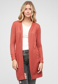 Lang, rustfarget cardigan i myk strikket stoff. Åpen frontdesign med to frontlommer og ribbede mansjetter. Båret over en hvit skjorte.