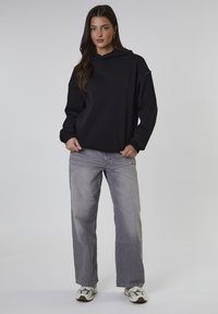 Felpa nera con cappuccio dal taglio comodo, abbinata a jeans grigi larghi. Il modello indossa sneakers bianche e grigie. Sfondo neutro.