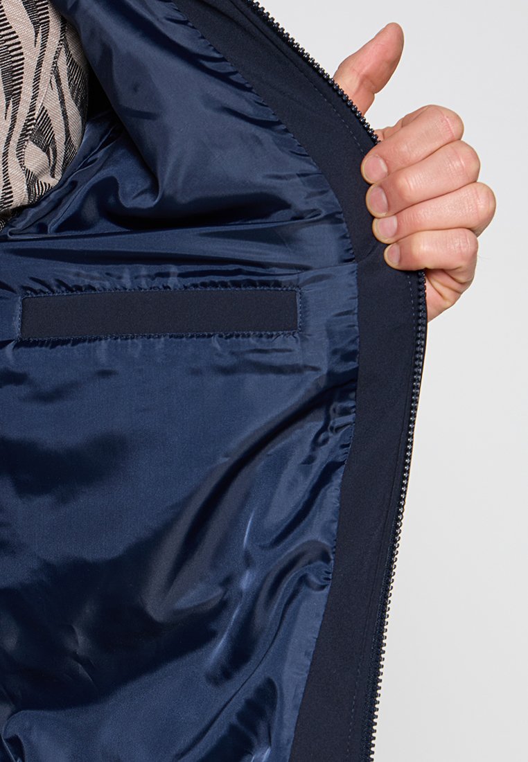 Una mano tiene aperto un cappotto blu navy che mostra liscio fodera interna con tasca interna rettangolare e bordo della cerniera nera.
