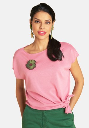 Frau mit dunklen Haaren in einem rosa Kurzarmtop und grünen Hosen, trägt eine grüne, blumenförmige Brosche und passende lange Blumenohrringe.