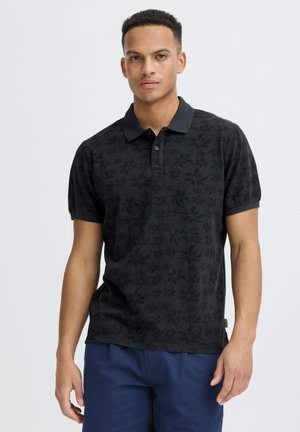 Hombre con una camisa polo negra con estampado floral y pantalones azules, de pie frente a un fondo claro y liso.