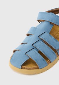 Ανοιχτό μπλε σανδάλια από μαλακό σουέντ, με πολλαπλά φαρδιά λουριά και επενδεδυμένη εσωτερική σόλα. Η σόλα είναι από μπεζ καουτσούκ με υφασμένη επιφάνεια.