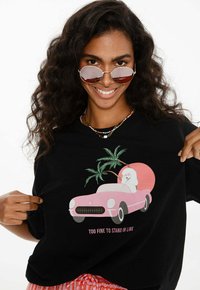 Usmívající se žena v kulatých slunečních brýlích a barevných náhrdelnících ukazuje na černé tričko s růžovým autem, palmami, psem a textem "Příliš fajn na to, abych stála ve frontě."