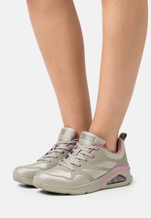 Jambes chaussées de baskets Skechers beige et rose avec lacets, tissu en maille, et semelles avec coussin d'air visible sur fond blanc.