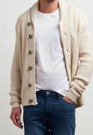 Man draagt een beige grofgebreid vest met grote knopen over een wit T-shirt en blauwe spijkerbroek, met één hand in de zak, tegen een effen achtergrond.