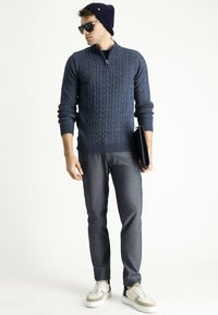 Dunkelblaues, kabelgroß gestricktes Sweatshirt mit hohem Kragen, kombiniert mit dunkelgrauen Hosen und weißen Sneakers mit grünen Akzenten. Das Model trägt eine Sonnenbrille und eine Mütze.