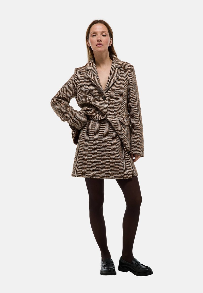 Blazer à motif pied-de-poule marron avec jupe assortie, présentant un col cranté, une fermeture à un bouton, et des poches avant, associé à des collants noirs et des chaussures noires.