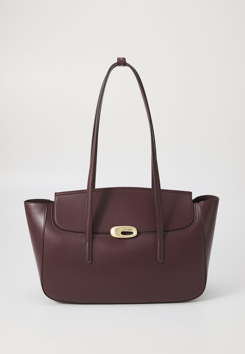 Sac à main en cuir bordeaux avec deux longues poignées, doté d'un fermoir oval en or et de détails de couture, au design structuré et à la texture lisse.