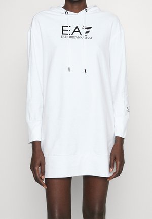 Persoon draagt een lange witte hoodie-jurk met zwart "EA7 Emporio Armani" logo op borst en mouw, staand tegen een effen achtergrond.