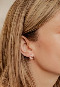 Ohrringe in Roségold mit floralem Design, die mit marcasit- und runden Zirkonia-Steinen verziert sind, vor einem sanft strukturierten Hintergrund.