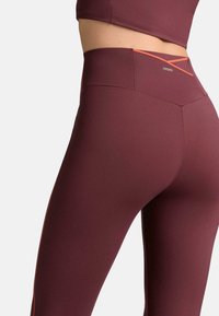 Bordové legíny s vysokým pásom, detailom na chrbte v tvare kríža a hladkou, textúrovanou látkou s oranžovými akcentmi pozdĺž strán.