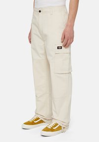Beige cargobroek van stevig materiaal, voorzien van een zijzak, verstevigde stiksels en een iets losse pasvorm. Gecombineerd met mosterdkleurige sneakers.