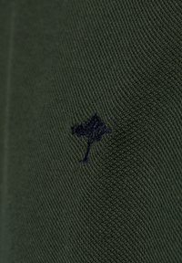 Mörkgrön tyg med en strukturerad väv, som har en liten marinblå trädlogotyp broderad nära nederkantens högra sida.