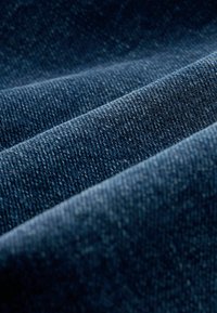 Sötétkék farmer anyag, jellegzetes textúrával. Az anyagon jól látható a diagonális anyagszerkezet és a hajtások mentén változó kék árnyalatok.