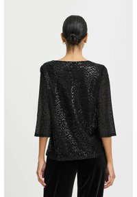Top noir transparent à manches trois-quarts, présentant un motif texturé et un léger éclat. Texture lisse avec une coupe ample et décontractée.