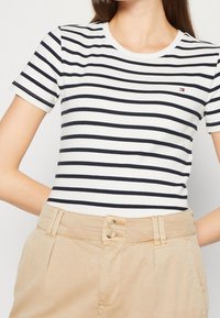 T-shirt blanc avec des rayures horizontales bleu marine, col rond et manches courtes. Associé à un pantalon beige avec une fermeture à double bouton.