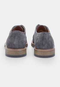 Scarpe in suede grigio con tacco basso, caratterizzate da una texture liscia, forma arrotondata e suola marrone a contrasto. Impunture a vista come accenti.