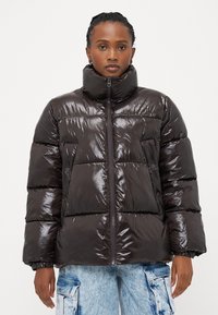Καφέ γυαλιστερό puffer μπουφάν με ψηλό γιακά, κλείσιμο με φερμουάρ και μπροστινές τσέπες. Διαθέτει τετράγωνο σχέδιο και ελαστικές μανσέτες.
