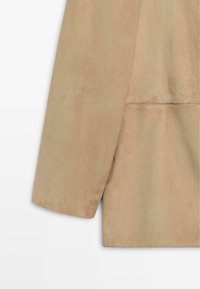 Manche de veste en daim beige et bas du dos avec couture visible et texture lisse sur un fond clair.
