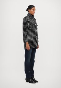 Abrigo a cuadros en blanco y negro con un cuello alto, que cuenta con dos bolsillos frontales y botones, combinado con jeans oscuros y botas negras hasta el tobillo.