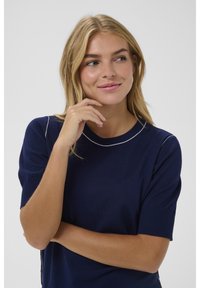 Šviesiaplaukė moteris, vilkinti tamsiai mėlyną trumpomis rankovėmis marškinėlį su balta apdaila, remiasi žandikauliu į delną, žiūri į šoną su nežymia šypsena, neutralus fonas.