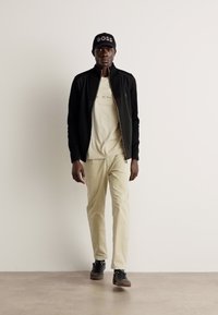 Jachetă neagră cu fermoar, tricou grafic bej, pantaloni bej deschis, adidași negri. Căciulă neagră cu "BOSS" brodată în alb.