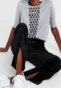 Grijs T-shirt met korte mouwen en zwart geometrisch patroon op de voorkant, gecombineerd met zwarte plooibroek met wijde pijpen en zijsplitten. Witte sneakers.