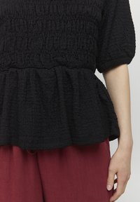 Sort tekstureret top med en samlet peplum-design og korte ærmer. Stoffet ser krøllet ud. Sammen med bordeaux bukser.