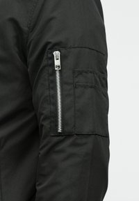Manche en tissu noir avec fermeture éclair verticale argentée et petite poche rectangulaire sur le haut du bras.