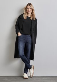 Svart dubbelknäppt kappa över en svart tröja, kombinerad med mörkblå jeans och vita sneakers med bruna detaljer på en grå bakgrund.