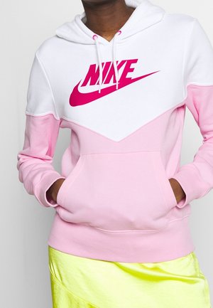 Personne portant un sweat à capuche Nike blanc et rose avec les mains dans la poche avant et une jupe jaune fluo.
