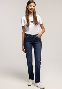 Młoda kobieta stoi z ręką w kieszeni, nosząc białą koszulkę z logo, ciemnoniebieskie dżinsy i białe trampki na jednolitym jasnym tle.
