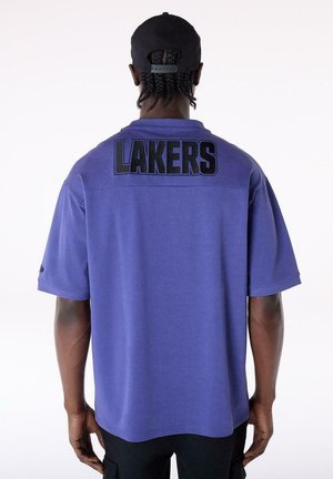 Person wearing a purple Lakers shirt with black text on the back and a black cap, shown from behind against a plain background.