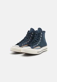 Høyhælte sneakers i marineblå semsket skinn med hvite gummisåler. Har sølvfargede snøringøyer og kontrasterende oransje detaljer. Klassisk tåhette-design.