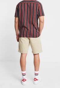 Korte mouwen, gestreept shirt in kastanjebruin, zwart en wit, gecombineerd met beige shorts. Witte sneakers met rode accenten en gestreepte sokken.