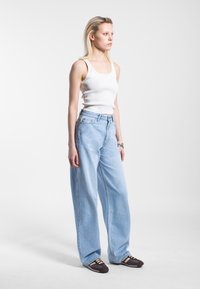 Lichtblauwe wijde denim jeans, gecombineerd met een witte geribde tanktop. Model draagt donkerbruine sneakers met accentkleuren.