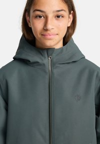 Giacca con cappuccio di colore verde scuro, realizzata in un tessuto liscio e leggero. Presenta una zip frontale e un logo ricamato sul petto.