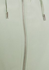 Jasnozielona kurtka na zamek z metalowym suwakiem i sznurkami. Gładka tkanina o subtelnym połysku, zaprojektowana z myślą o komforcie i stylu.