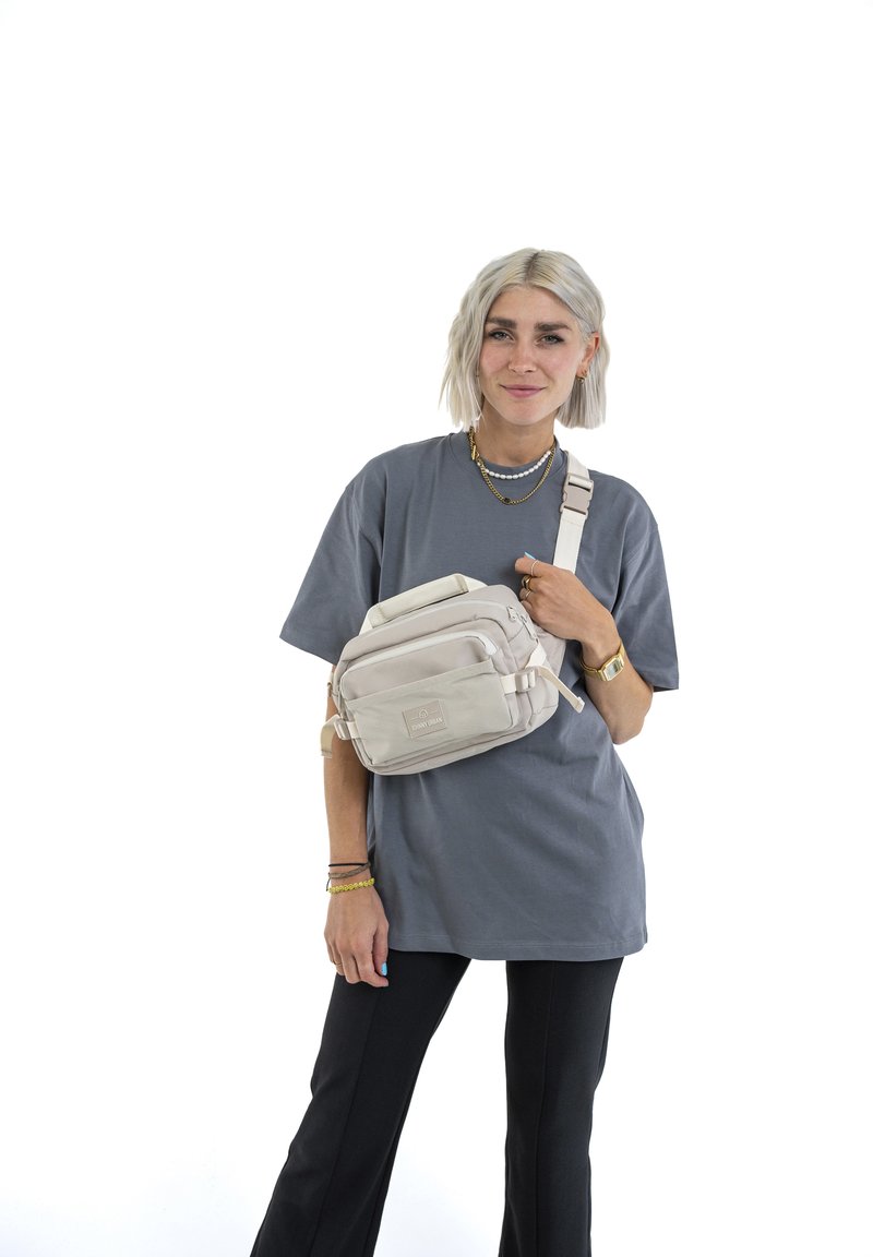 Borsa a tracolla beige con più scomparti, tracolla regolabile e etichetta patch frontale. Indossata da una persona con una t-shirt grigia e pantaloni neri.