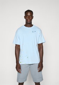 Hele sinine puuvillane T-särk, millel on rinnal sinine Nike logo ja värviline pritsmetega muster allservas. Sobib hallide lühikeste pükstega.