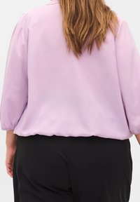 Blusa lavanda con maniche a tre quarti, collo rotondo e polsini elasticizzati. Tessuto liscio con una morbida lucentezza, abbinata a pantaloni neri.